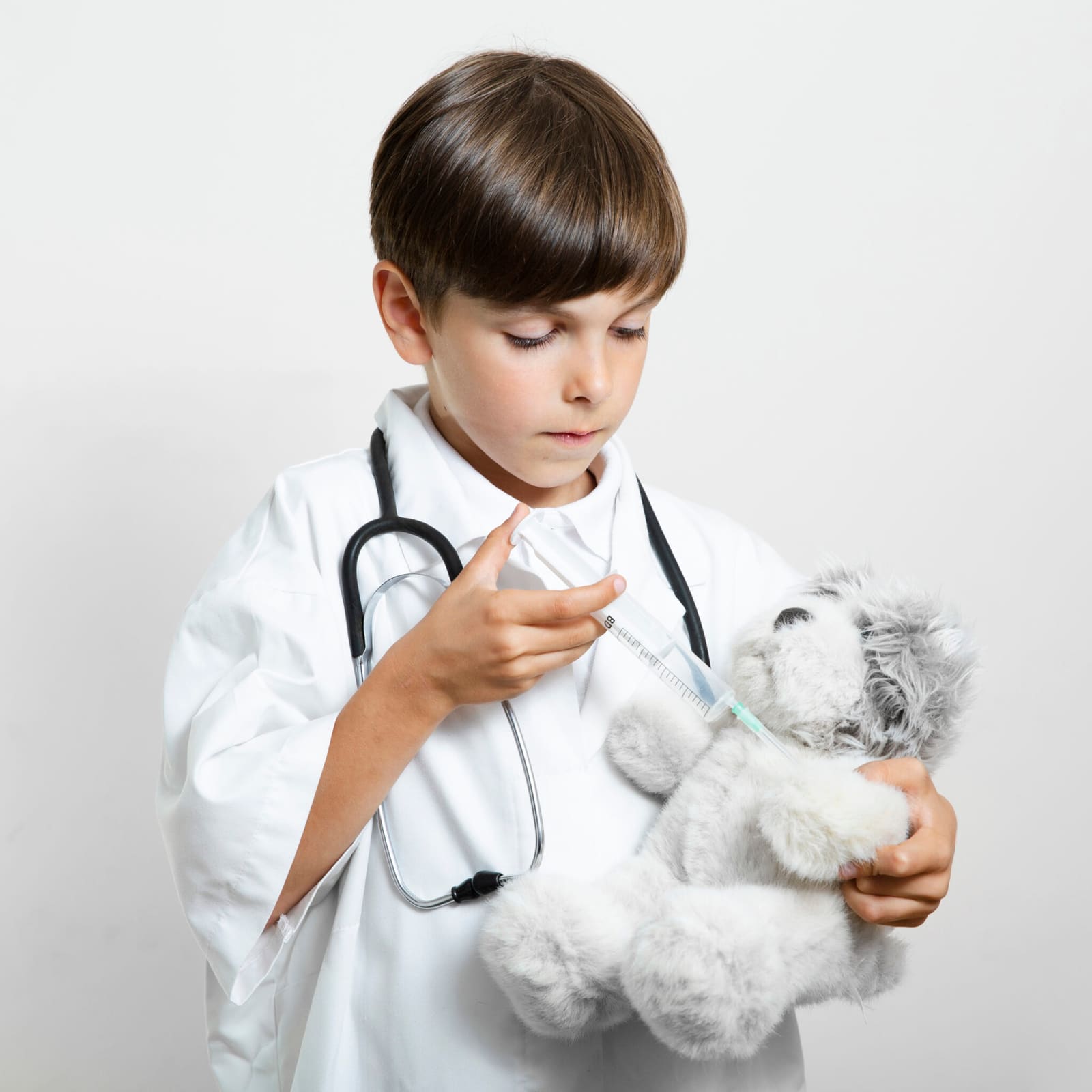 Menino vestido de médico, com jaleco e estetoscópio, brincando de aplicar uma injeção em um urso de pelúcia